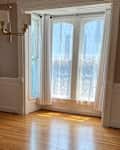 A bright room with hardwood floors, large windows with sheer curtains, and a modern chandelier.