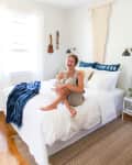 Woman sitting on a white bed with blue pillows, a blue throw, and wall decor in a bright bedroom.
