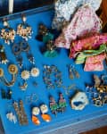 Assorted colorful earrings and jewelry displayed on a blue tray next to patterned fabric pouches and a pink case.
