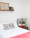 Cozy bedroom with white bedding, colorful pillow, red throw, wall art, shelf with books, and a potted plant on a side table.