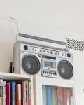 Vintage boombox on a white shelf above colorful books in a bright room.