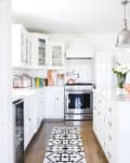White kitchen with stainless steel appliances, pendant lights, patterned rug, and a vase with flowers on the island.