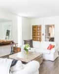 Bright living room with white sofas, wooden coffee table, large mirrors, and green plants on a console table.
