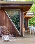 Wooden deck with a white wire chair, outdoor dining table set, and string lights, adjacent to a modern house with large windows.