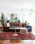 Living room with brown leather sofa, colorful pillows, red patterned rug, wicker chair, and large plant by the window.