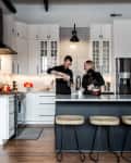 Couple preparing drinks in a modern kitchen with white cabinets, black island, and wooden stools.