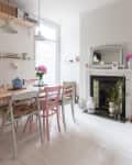 Dining room with pastel chairs, wooden table, blue teapot, floral cushions, and a decorative fireplace with plants.