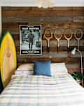 Bedroom with wood accent wall, vintage tennis rackets, Taxi Driver poster, surfboard, and plaid bedspread.