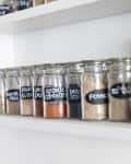 Glass jars with black labels filled with various spices on a white shelf.