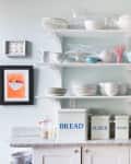 Open kitchen shelves with assorted mugs, bowls, glasses, and jars labeled bread, flour, and sugar on a granite countertop.