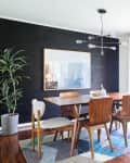 Mid-century modern dining room with wooden table, mixed chairs, geometric rug, black accent wall, and large plant.