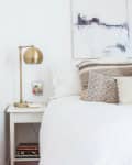 A white bed with patterned pillows next to a white side table with a funky brass table lamp.