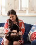 Woman in floral dress holding a black cat on a blue sofa with colorful pillows and framed artwork on the wall.