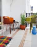 Small balcony with colorful chairs, striped rug, potted plants, and a blue vase, overlooking a cityscape.