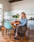 Woman sitting at a wooden dining table in a bright kitchen with large windows, plants, and light blue cabinets.