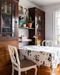 Cozy dining nook with floral tablecloth, wooden cabinets, potted plants, and a colorful window decoration.