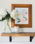Wooden shelf with a glass vase of white hydrangeas, a lit candle, and a framed floral painting.