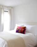 Bright bedroom with white bedding, colorful pillows, sheer curtains, and a woven basket near the window.