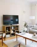 Living room with gray sofa, wooden coffee table, TV on stand, and white curtains. Decor includes candles and flowers.