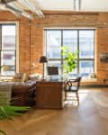 Loft-style office with exposed brick walls, large windows, leather sofa, wooden desk, and dining table with chairs.