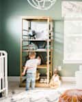 Child reaching for toys on a wooden shelf in a green nursery with a white armchair, lamp, and changing table.