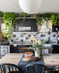 Cozy kitchen with black cabinets, patterned backsplash, hanging plants, and a rustic wooden table set for a meal.