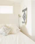 A light bedroom with white bedding, fuzzy pillows, a window, and a black-and-white patterned wall hanging beside the bed.