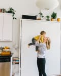 Woman lifting child in a bright kitchen with white cabinets, books, and plants.