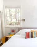Bright bedroom with a white bed, colorful pillows, wall lamps, and two windows overlooking greenery.
