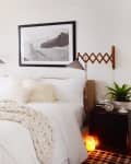 Cozy bedroom with white bedding, chunky knit throw, wall sconces, plants, and a framed black-and-white landscape photo.