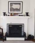 White brick fireplace with black screen, framed sheep art, decorative vases, and a wooden chair in a living room.