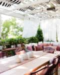Outdoor patio with white pergola, sectional sofa with pink cushions, striped rug, and dining table surrounded by greenery.