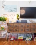Wooden sideboard with drawers, plants, and decor under a wall-mounted TV, surrounded by toys and books in wire baskets.