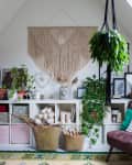 Cozy attic room with macramé wall hanging, plants, a brown chair, and a white shelf with storage baskets and decor.