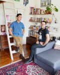 Two people in a cozy living room with bookshelves, a gray sofa, colorful rug, and wall art.