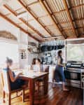 Rustic kitchen with corrugated metal ceiling, wooden floor, two women at a table, and one cooking by the stove.