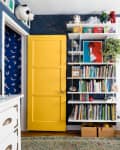 Colorful children's room with a yellow door, blue walls, white shelves filled with books, toys, and a white chair.