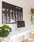 Home office with a large wall calendar, white desk, computer, plants, and wall art.