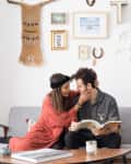 Couple sitting on a gray sofa, woman in red dress, man reading a book, surrounded by eclectic wall art and decor.