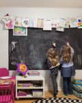Two children drawing on a large chalkboard in a colorful playroom with books and artwork displayed.