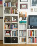 Bookshelf filled with colorful books, magazines, and decor items next to a TV, with framed art on the wall.