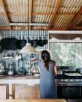 Rustic kitchen with corrugated metal walls, hanging pots, wooden shelves, and a woman cooking at the stove.