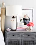 Bedside table with a white lamp, framed photo, pink flower, decorative bicycle, and books next to a white bed.