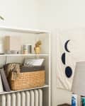 White shelf with books, basket, and decor items next to a lamp and wall art with moon phases.