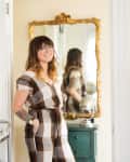 Woman in checkered jumpsuit standing by a bar cart with glasses and copper mugs, next to a gold-framed mirror.