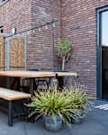 Outdoor patio with wooden picnic table, black metal chairs, potted plants, and string lights against a brick wall.