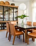 Dining room with wooden table, six chairs, glass cabinet, and large plant vase near sheer curtained windows.