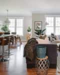 Open-concept living room and kitchen with gray sofa, wooden bar stools, large windows, and decorative plants.