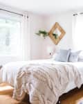 Bright bedroom with a white bed, knitted throw, wooden nightstand, and wall decor. Basket with pillows on the floor.