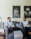 Two men sitting on a gray sofa in a living room with owl artwork and a wooden coffee table.